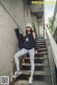 A woman in a black top and white leggings leaning against a wall.