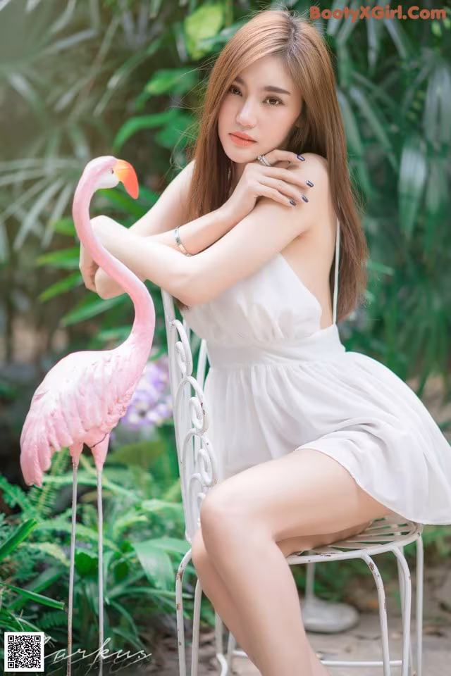 A woman in a white dress sitting on a white chair next to a pink flamingo.