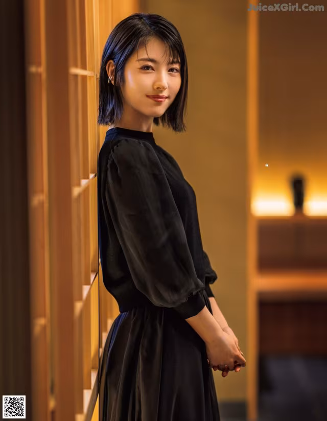 A woman in a black dress leaning against a wall.